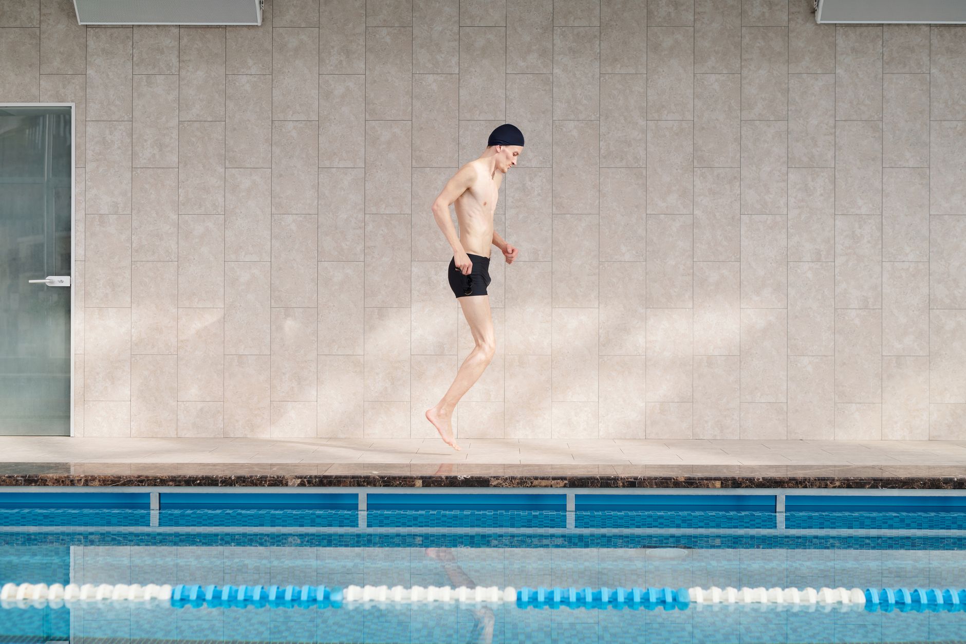 Amputee man jumping poolside during morning workout in indoor swimming pool.