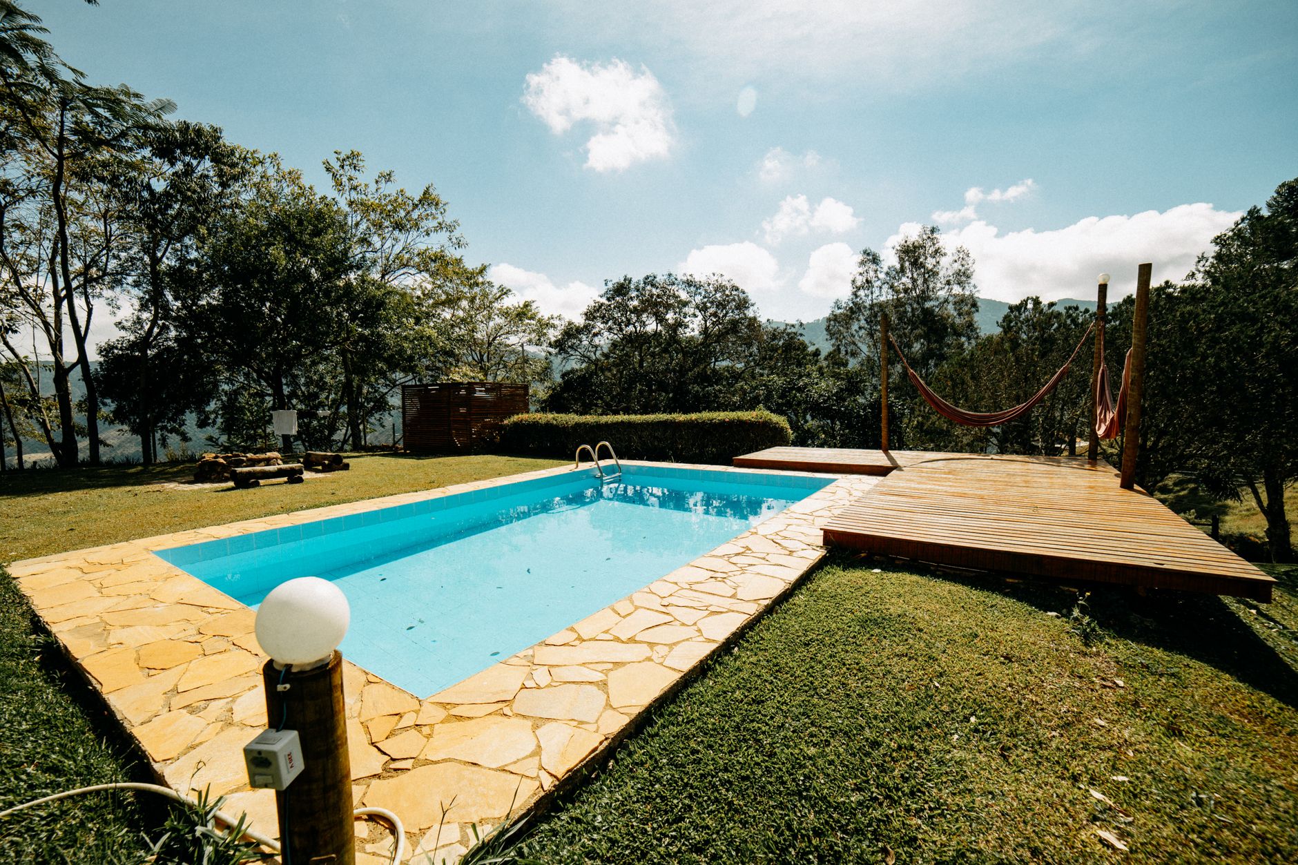 A serene outdoor pool with a wooden deck and hammock surrounded by lush greenery on a sunny day.
