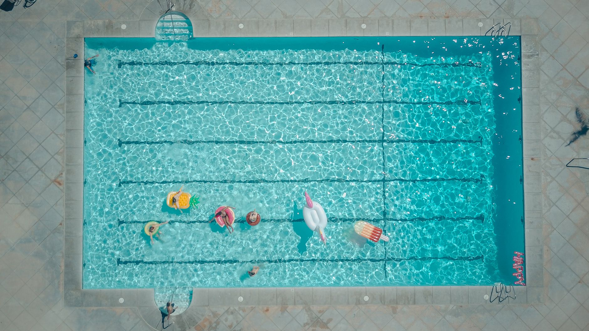 Top-down view of a swimming pool with colorful floats and people enjoying a sunny pool party.